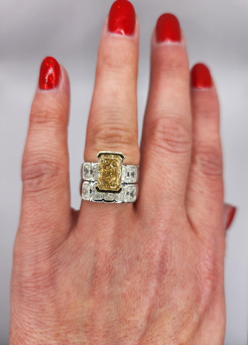 May include: A close-up of a hand wearing a ring with a large, rectangular, yellow diamond surrounded by smaller diamonds. The ring is set in white gold and has a band of smaller diamonds.