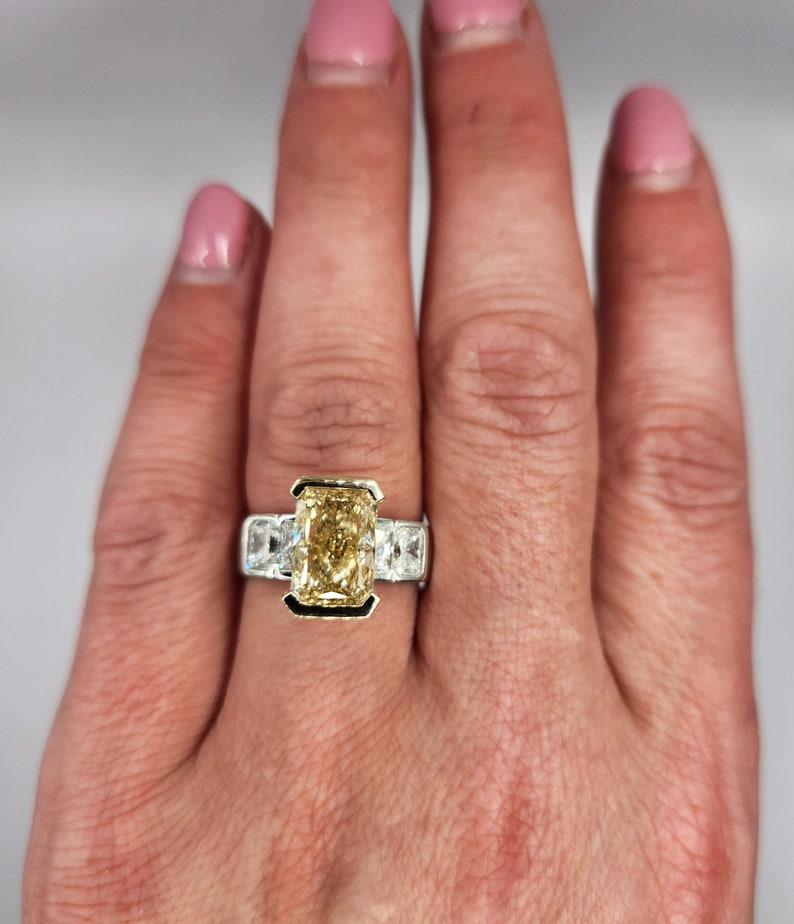 May include: A gold and white engagement ring with a large, rectangular, yellow diamond in the center. The ring is set with smaller diamonds on either side of the center stone.