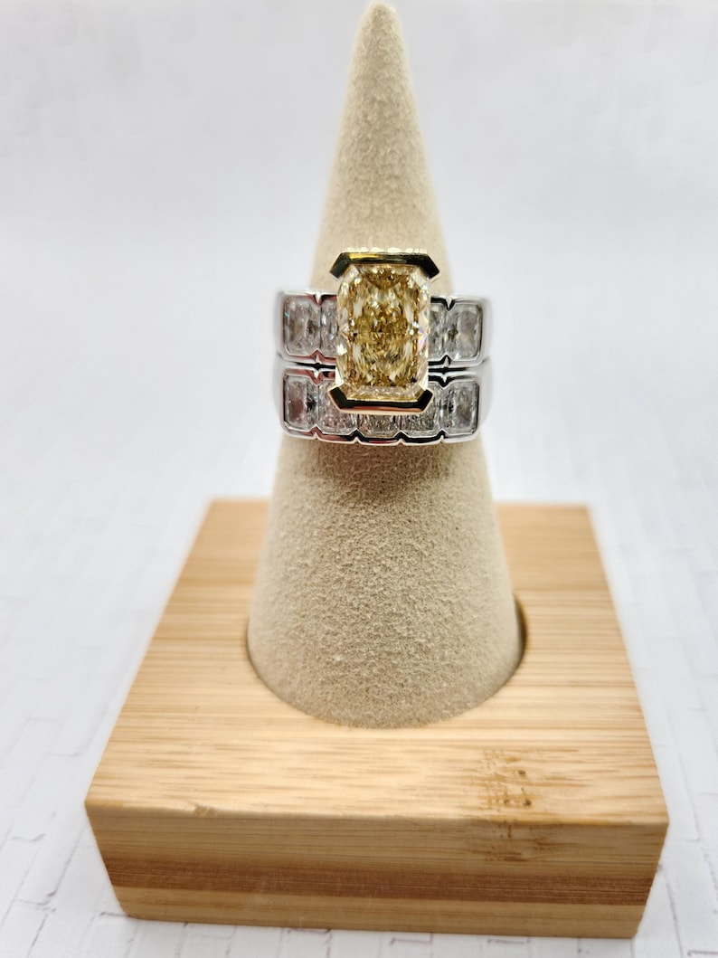 May include: A yellow diamond engagement ring with a square-shaped center stone and a band of smaller diamonds. The ring is set on a white cone-shaped display stand.