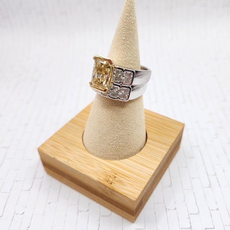 May include: A yellow diamond engagement ring with a square cut stone set in a white gold band. The ring is on a wooden ring display stand.