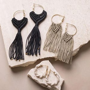 May include: A selection of handmade macrame earrings. The earrings have gold-tone hoops and macrame detailing in black, beige, and white. The black and beige earrings have long fringe tassels, while the white earrings feature a looped design.