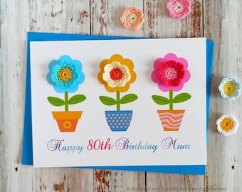 Op de afbeelding: Een witte kaart met een blauwe rand en drie kleurrijke bloemen in potten. De kaart zegt "Happy 80th Birthday Mum".