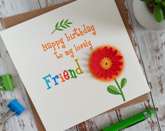 May include: A white card with the text "Happy birthday to my lovely Friend" in rainbow colors. A red crocheted flower with a yellow center is on the card.