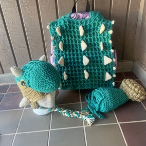 May include: A green crocheted dinosaur backpack, hat, and tail. The backpack has white spikes and a waffle stitch pattern. The hat has a brown snout and white horns. The tail is a long, crocheted tube with a brown tip.