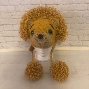 May include: A crocheted lion hat with a yellow mane, brown face, and green eyes. The hat has two yellow pom-poms on the ends of the ear flaps.