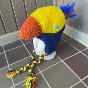 May include: A crocheted toucan hat with a yellow, orange, and blue color scheme. The hat has a long braided tail with tassels.