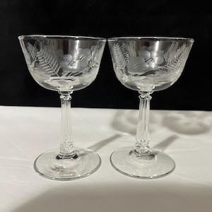 May include: Two clear champagne glasses with etched floral and fern designs. The glasses have a wide bowl, a long stem, and a round base. The etched design is visible on the bowl of each glass. The glasses are placed on a white surface.