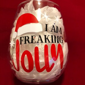 Pode incluir: Uma ta&ccedil;a de vinho de vidro transparente com um design de chap&eacute;u de Papai Noel vermelho e branco e o texto "I am freaking jolly" em letras pretas.