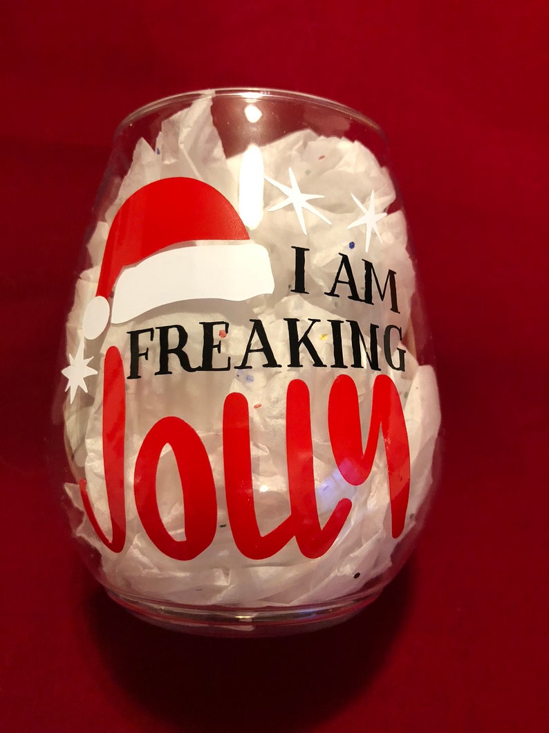 Pode incluir: Uma ta&ccedil;a de vinho de vidro transparente com um design de chap&eacute;u de Papai Noel vermelho e branco e o texto "I am freaking jolly" em letras pretas.