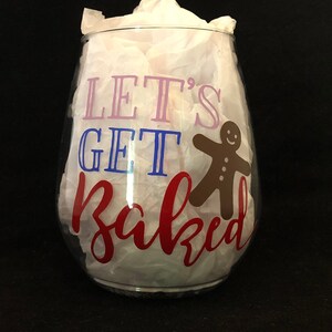 Pode incluir: Uma ta&ccedil;a de vinho de vidro transparente com o texto "Let's Get Baked" em letras rosa, azul e vermelho. Um homem de gengibre est&aacute; &agrave; direita do texto.