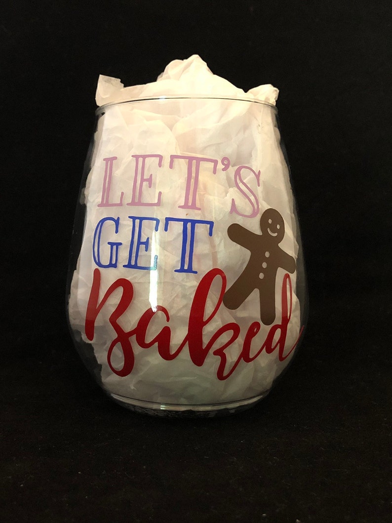 Pode incluir: Uma ta&ccedil;a de vinho de vidro transparente com o texto "Let's Get Baked" em letras rosa, azul e vermelho. Um homem de gengibre est&aacute; &agrave; direita do texto.