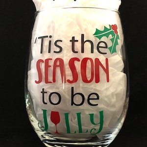 Pode incluir: Uma ta&ccedil;a de vinho de vidro transparente com o texto 'Tis the season to be jolly' em letras pretas, vermelhas e verdes. Uma pequena baga vermelha e uma folha de azevinho verde est&atilde;o no lado direito do copo.