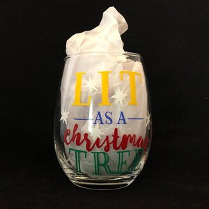 Pode incluir: Uma ta&ccedil;a de vinho de vidro transparente com o texto "LIT AS A CHRISTMAS TREE" em letras amarelas, vermelhas, azuis e verdes com estrelas brancas.