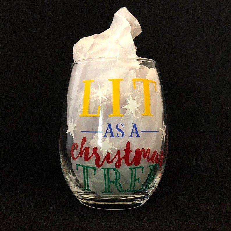 Pode incluir: Uma ta&ccedil;a de vinho de vidro transparente com o texto "LIT AS A CHRISTMAS TREE" em letras amarelas, vermelhas, azuis e verdes com estrelas brancas.