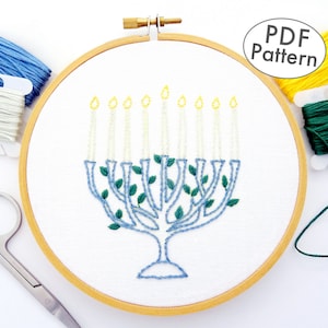 May include: A PDF pattern for a blue and green embroidered menorah with yellow candles. The pattern is displayed in a wooden embroidery hoop with white fabric. The image also shows blue, white, yellow, and green embroidery floss.