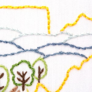 Op de afbeelding: Close-up van een handgestikt borduurmotief met een landschap met een gele lucht, blauw water en groene bomen. Het motief is geborduurd op witte stof.