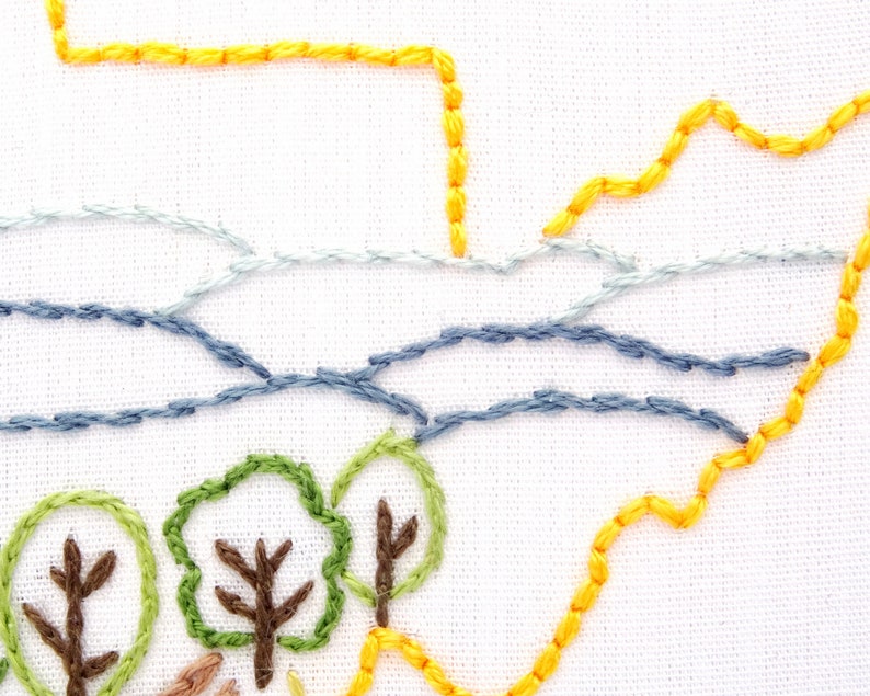 Op de afbeelding: Close-up van een handgestikt borduurmotief met een landschap met een gele lucht, blauw water en groene bomen. Het motief is geborduurd op witte stof.