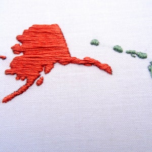 K&ouml;nnte beinhalten: Gestickte Karte von Alaska in rotem Faden und Hawaii in gr&uuml;nem Faden auf wei&szlig;em Stoffgrund.