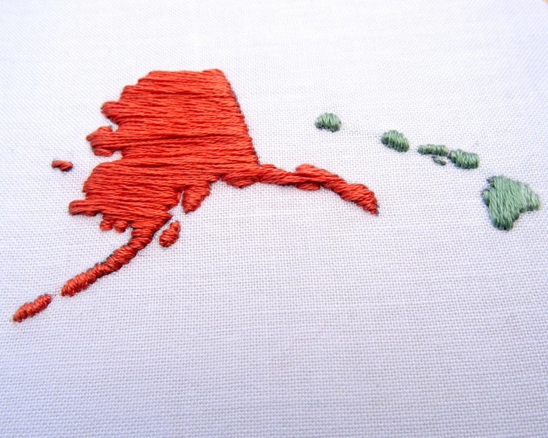 K&ouml;nnte beinhalten: Gestickte Karte von Alaska in rotem Faden und Hawaii in gr&uuml;nem Faden auf wei&szlig;em Stoffgrund.