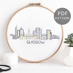 May include: Embroidered cityscape of Glasgow, Scotland, in a wooden embroidery hoop. The skyline is stitched in blue and gold thread on white fabric. The words "PDF PATTERN" are in a grey circle in the top right corner.