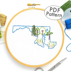 Könnte beinhalten: Stickerei-Design des Bundesstaates Maryland mit Bäumen und Gras in grünem, blauem und braunem Garn auf weißem Stoff in einem hölzernen Stickrahmen. Der Text "PDF Pattern" befindet sich in der oberen rechten Ecke des Bildes.
