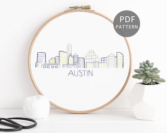 Puede incluir: Un aro de bordado blanco con un horizonte bordado en azul y amarillo de Austin, Texas. El texto "AUSTIN" está debajo del horizonte. El aro está sobre una superficie blanca con una bola de hilo blanco y unas tijeras negras.