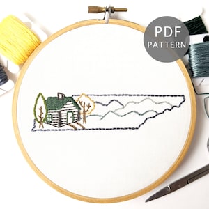 May include: Embroidered design of the state of Tennessee with a cabin, trees, and mountains. The design is stitched on white fabric in a wooden embroidery hoop. The hoop is surrounded by embroidery floss in various colors.