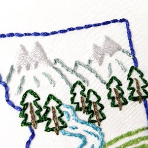 May include: Embroidered design of a mountain scene with blue, green, and white thread. The design features three mountains, trees, and a river.