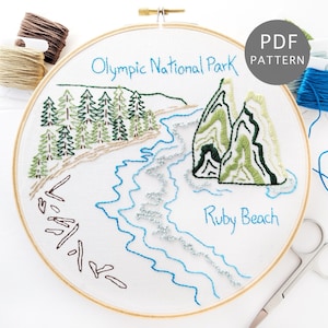 May include: Embroidery hoop with a design of a beach scene with trees, rocks, and waves. The text "Olympic National Park" and "Ruby Beach" are embroidered on the hoop. The hoop is surrounded by embroidery floss in various colors.