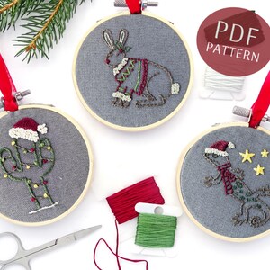 May include: Three embroidery hoop ornaments with Christmas themes. The ornaments feature a cactus wearing a Santa hat, a lizard wearing a Santa hat and scarf, and a bunny wearing a sweater. The ornaments are on a white surface with red and green embroidery floss.