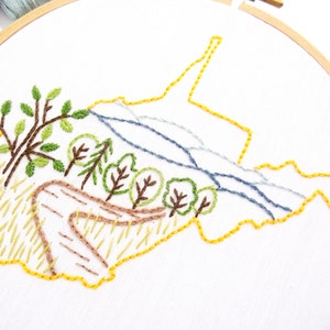 Op de afbeelding: Borduurwerk van een staatsgrens met een landschapsscene erin. De staat is omlijnd met gele draad, en het landschap toont bomen, een kronkelend pad en een rivier. Het borduurwerk is uitgevoerd op witte stof.