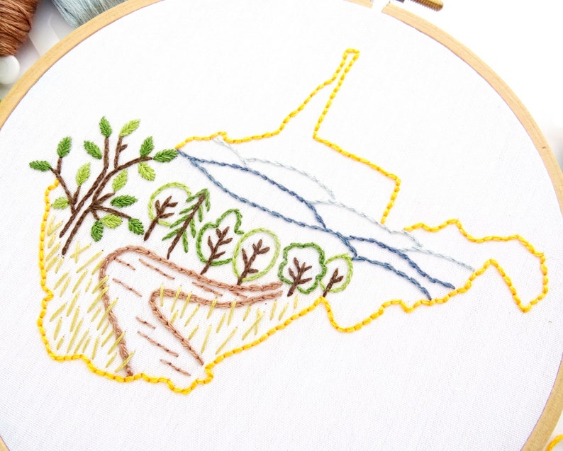 Op de afbeelding: Borduurwerk van een staatsgrens met een landschapsscene erin. De staat is omlijnd met gele draad, en het landschap toont bomen, een kronkelend pad en een rivier. Het borduurwerk is uitgevoerd op witte stof.