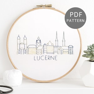Puede incluir: Bordado en bastidor con el horizonte de Lucerna en hilo azul marino y dorado. El diseño está cosido sobre tela blanca y enmarcado en un aro de madera. La palabra "LUCERNE" está bordada debajo. Un círculo gris con el texto "PDF PATTERN" está en la parte superior derecha.