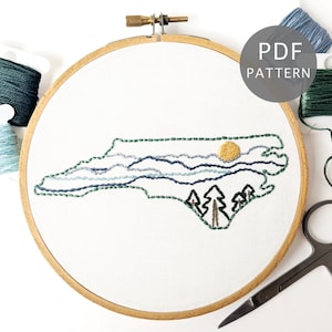 May include: Embroidered hoop art featuring a stylized outline of the state of North Carolina with a sun, mountains, and trees. The hoop is surrounded by embroidery floss in shades of green and blue.