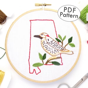 Puede incluir: Arte de bordado en aro con un pájaro posado en una rama, delineado en rojo, que se asemeja al estado de Alabama. El aro está rodeado de hilo de bordar en varios colores, incluyendo verde, marrón, amarillo y rojo.