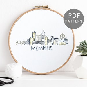 May include: A white embroidery hoop with a black and yellow stitched skyline of Memphis, Tennessee. The words "MEMPHIS" are stitched in black below the skyline. The hoop is on a white surface with a ball of white yarn and a pair of black scissors.