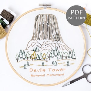 May include: An embroidery hoop featuring a stitched design of Devils Tower National Monument. The design shows the tower in brown and grey, with green trees and the text "Devils Tower National Monument". The words "PDF PATTERN" are also visible.