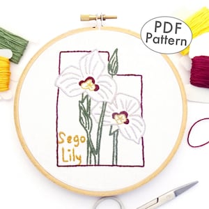 Può includere: Un cerchio da ricamo bianco con un disegno di un fiore di Sego Lily in filo bianco, verde e bordeaux. Il testo "Sego Lily" è ricamato in filo dorato. Il cerchio è circondato da filo da ricamo in giallo, verde e bordeaux.