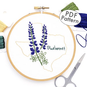 May include: Embroidered hoop art featuring a bluebonnet flower design in the shape of the state of Texas. The hoop is surrounded by embroidery floss in various colours, including blue, green, and yellow.