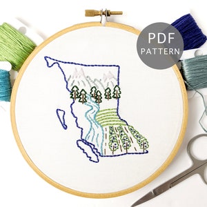 May include: Embroidered hoop art featuring a stylized outline of the province of British Columbia, Canada, with mountains, trees, and a river. The hoop is surrounded by embroidery floss in various shades of green and blue.