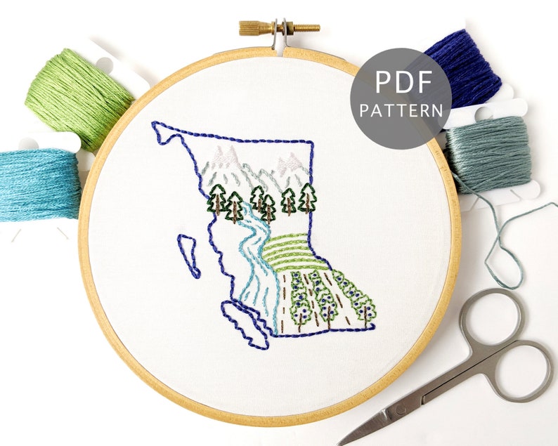 May include: Embroidered hoop art featuring a stylized outline of the province of British Columbia, Canada, with mountains, trees, and a river. The hoop is surrounded by embroidery floss in various shades of green and blue.