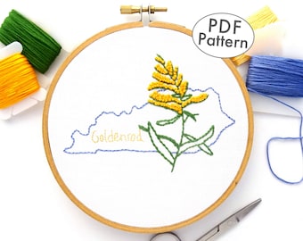 Könnte beinhalten: Eine gestickte Darstellung des Bundesstaates Kentucky mit einer Goldrute und dem Wort "Goldenrod", das mit gelbem Garn gestickt ist. Das Design befindet sich auf weißem Stoff in einem hölzernen Stickrahmen. Es gibt Spulen mit gelbem, grünem und blauem Stickgarn.