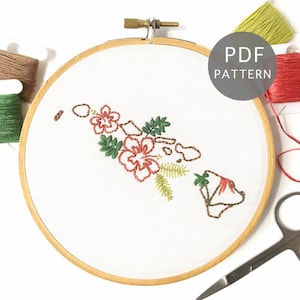 May include: Embroidery hoop with a white fabric depicting the outline of the Hawaiian islands with red hibiscus flowers and green leaves. The hoop is made of wood and has a brown finish. There are three spools of embroidery floss in the image: one green, one brown, and one red.