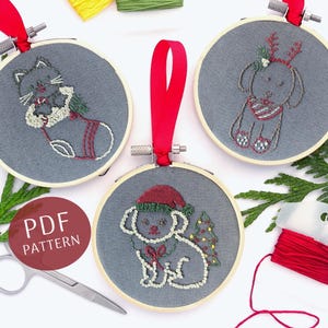 May include: Three embroidered Christmas ornaments featuring a cat wearing a stocking, a dog wearing a reindeer headband, and a dog wearing a Santa hat. The ornaments are on a white background with green foliage and red ribbon.
