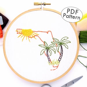 May include: Embroidery hoop with a white fabric showing an embroidered outline of the state of Florida with a sun and two palm trees. The hoop is surrounded by embroidery floss in various colors.