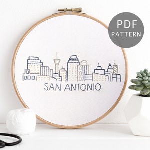 Puede incluir: Arte de bordado en aro con el horizonte de San Antonio, Texas. El diseño está hecho con hilo azul y amarillo sobre tela blanca. El aro es de madera y tiene un acabado natural.