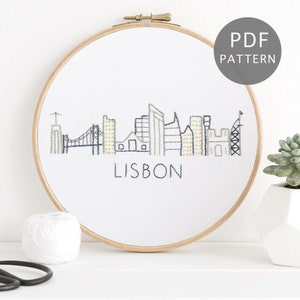May include: A white embroidery hoop with a black and white embroidered cityscape of Lisbon, Portugal. The text "LISBON" is embroidered below the cityscape. The hoop is on a white surface with a ball of white yarn and a pair of black scissors.