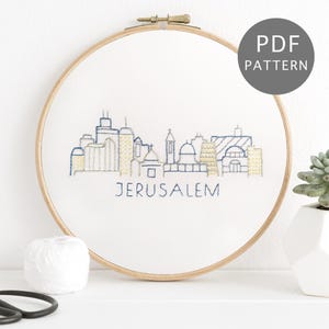 May include: An embroidered hoop art piece with a skyline design and the word "JERUSALEM" stitched in blue thread. The buildings are in blue and yellow. A PDF pattern is available.