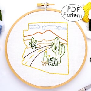 May include: A white embroidery hoop with a yellow outline, featuring a hand-stitched design of the state of Arizona with a desert landscape, including mountains, a road, and cacti. The text "PDF Pattern" is visible in the upper right corner of the image.