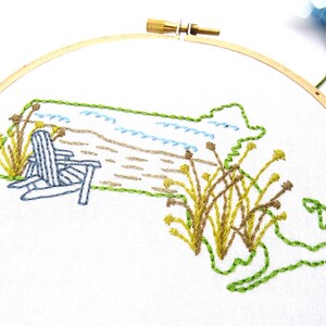 Puede incluir: Dise&ntilde;o bordado del estado de Massachusetts con una escena de playa, que incluye una silla Adirondack azul, arena marr&oacute;n, olas azules y juncos verdes y amarillos.